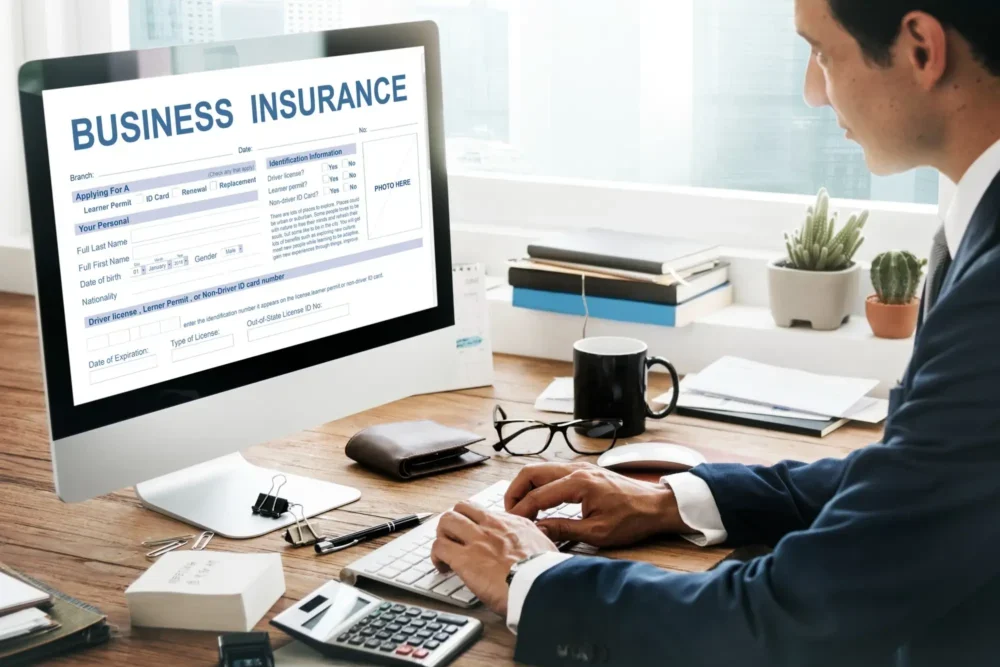 A person in a suit fills out a business insurance form on a desktop computer, surrounded by office supplies and a plant.